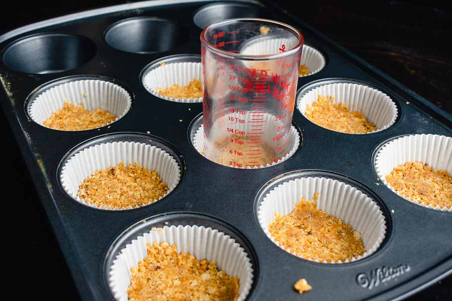 Pressing the graham cracker crumb mixture into the cupcake liners using a measuring glass